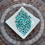 A square, white glass dish with a teal dot design. The dots vary in size and are clustered in the center of the dish. The dish is on a mosaic tile table with a circular pattern of brown, gray, and white tiles.