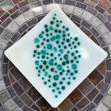 A square, white glass dish with a teal dot design. The dots vary in size and are clustered in the center of the dish. The dish is on a mosaic tile table with a circular pattern of brown, gray, and white tiles.
