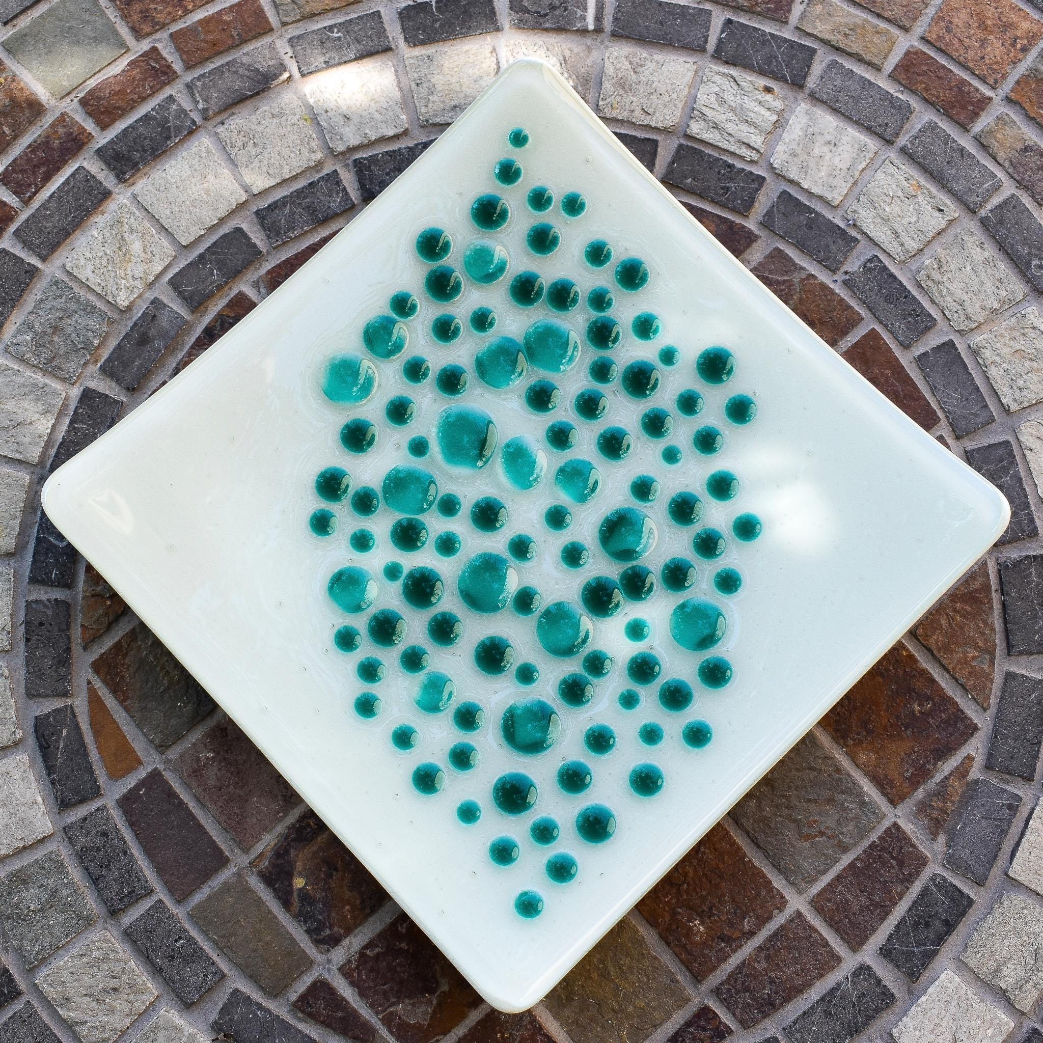 A square, white glass dish with a teal dot design. The dots vary in size and are clustered in the center of the dish. The dish is on a mosaic tile table with a circular pattern of brown, gray, and white tiles.