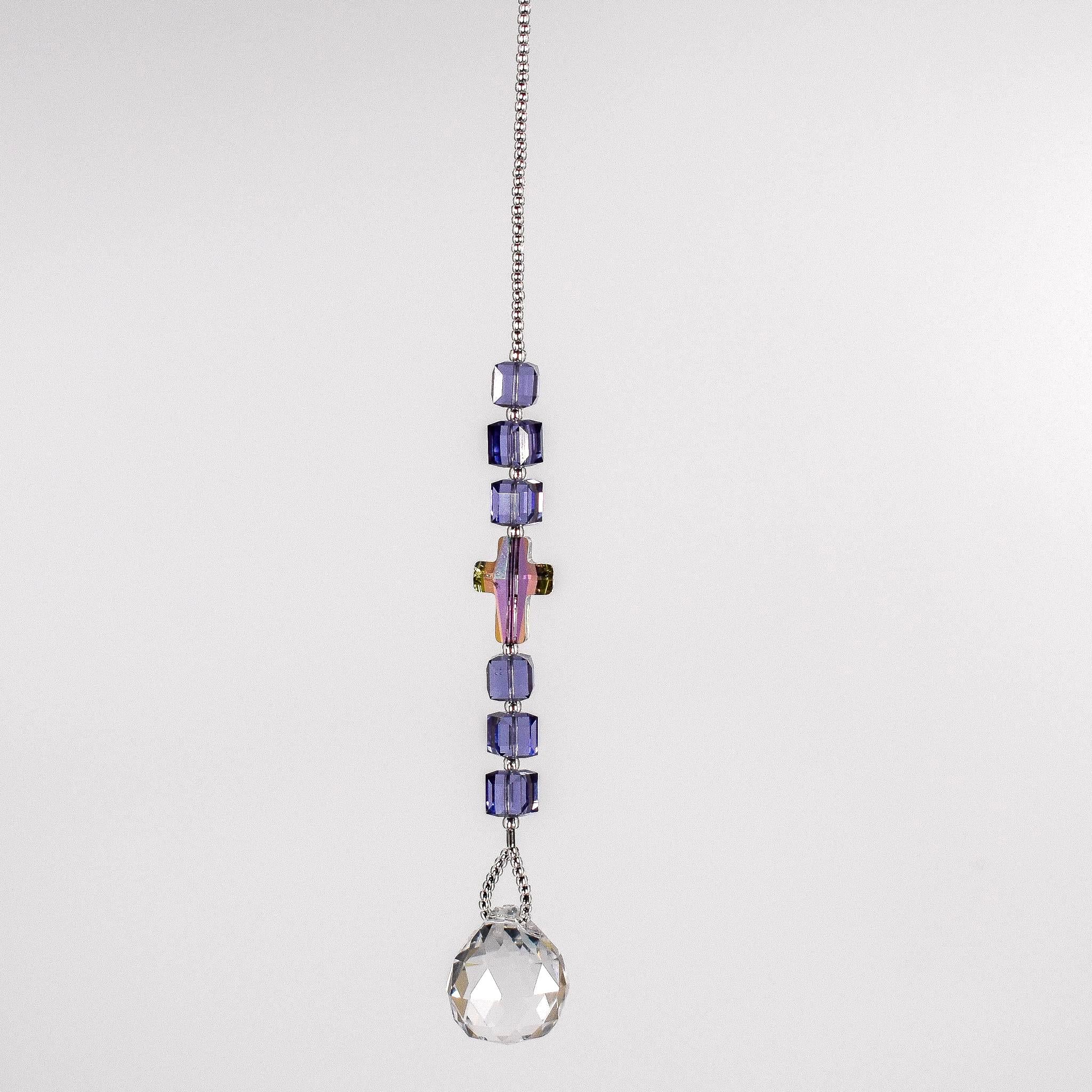 Small sun catcher with six purple crystal cube beads, a single crystal cross in paradise shine color, anchored by glass crystal prism. Hanging in front of white background.