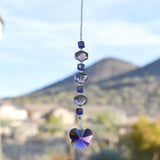 A handmade sun catcher featuring purple and iridescent crystal beads, finished with a faceted crystal butterfly pendant that reflects light in purple, blue, and gold tones. Suspended from a wire covered in silver-lined seed beads with a desert mountain in the background.
