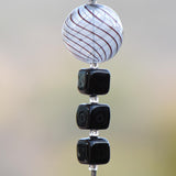Large, round, acrylic bead that is clear with brown swirls beaded on wire and hanging vertically, Below the round bead are 3 black cubic beads made of Murano glass. Background is blurred.