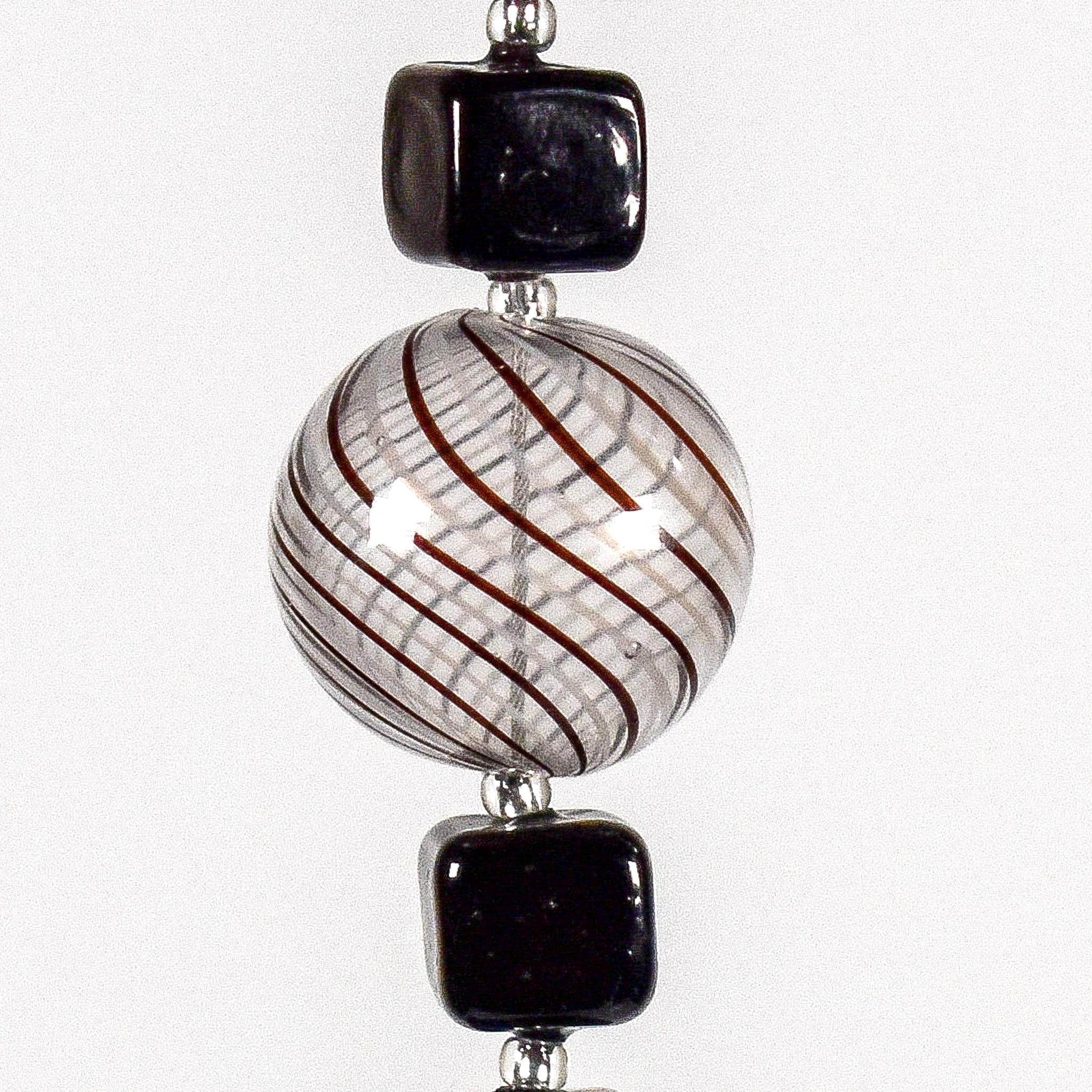 Large, round, acrylic bead that is clear with brown swirls beaded on wire and hanging vertically, flanked by 2 black cubic beads made of Murano glass. White background.