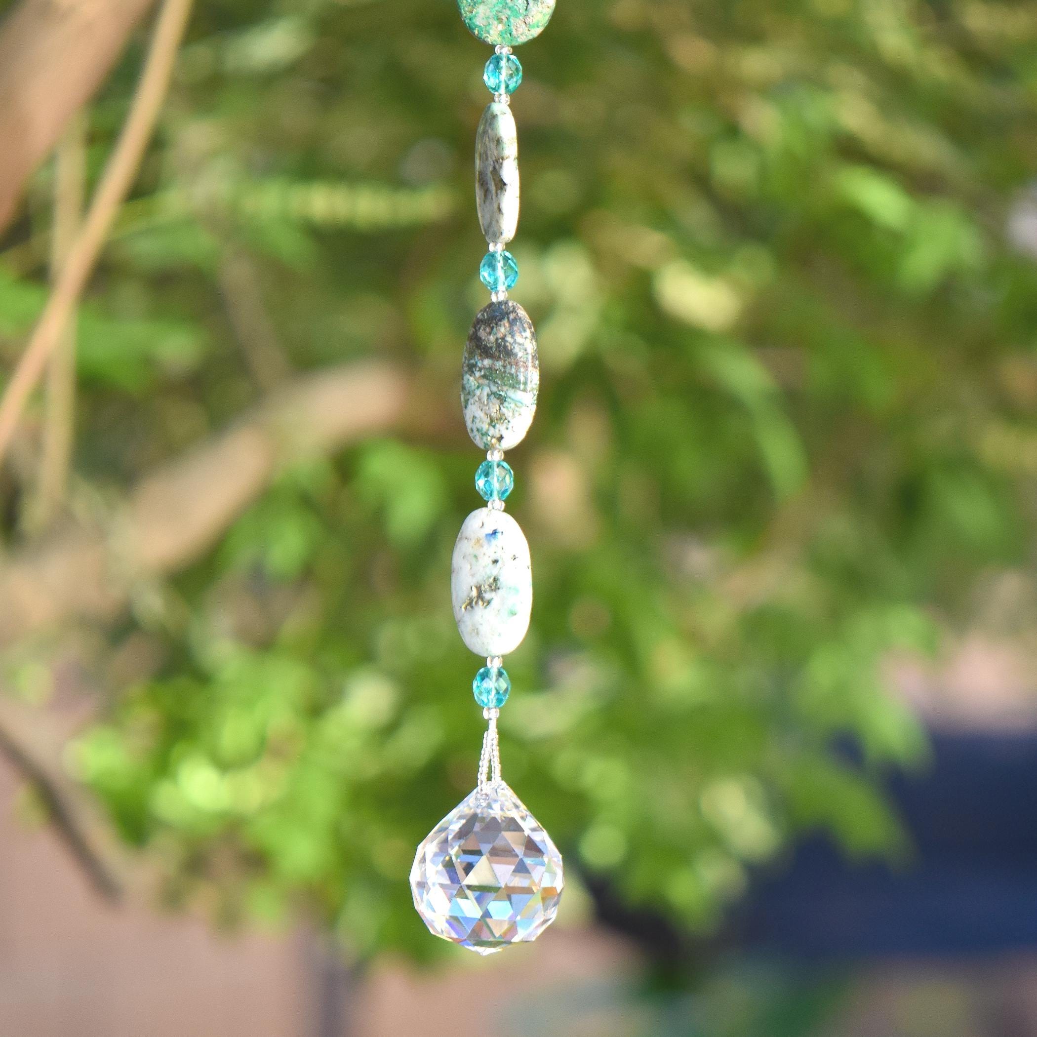 The bottom of a long hanging sun catcher made of large oval azurite malachite stone beads paired with small turquoise glass beads and a large crystal prism. This photo shows the bottom 3 stone beads, 3 glass beads and the prism, against a blurred foliage background.