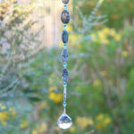 Long hanging sun catcher made of large oval azurite malachite stone beads paired with small turquoise glass beads and a large crystal prism. This photo shows the bottom 5 stone beads, 6 glass beads and the prism, against a blurred foliage background.