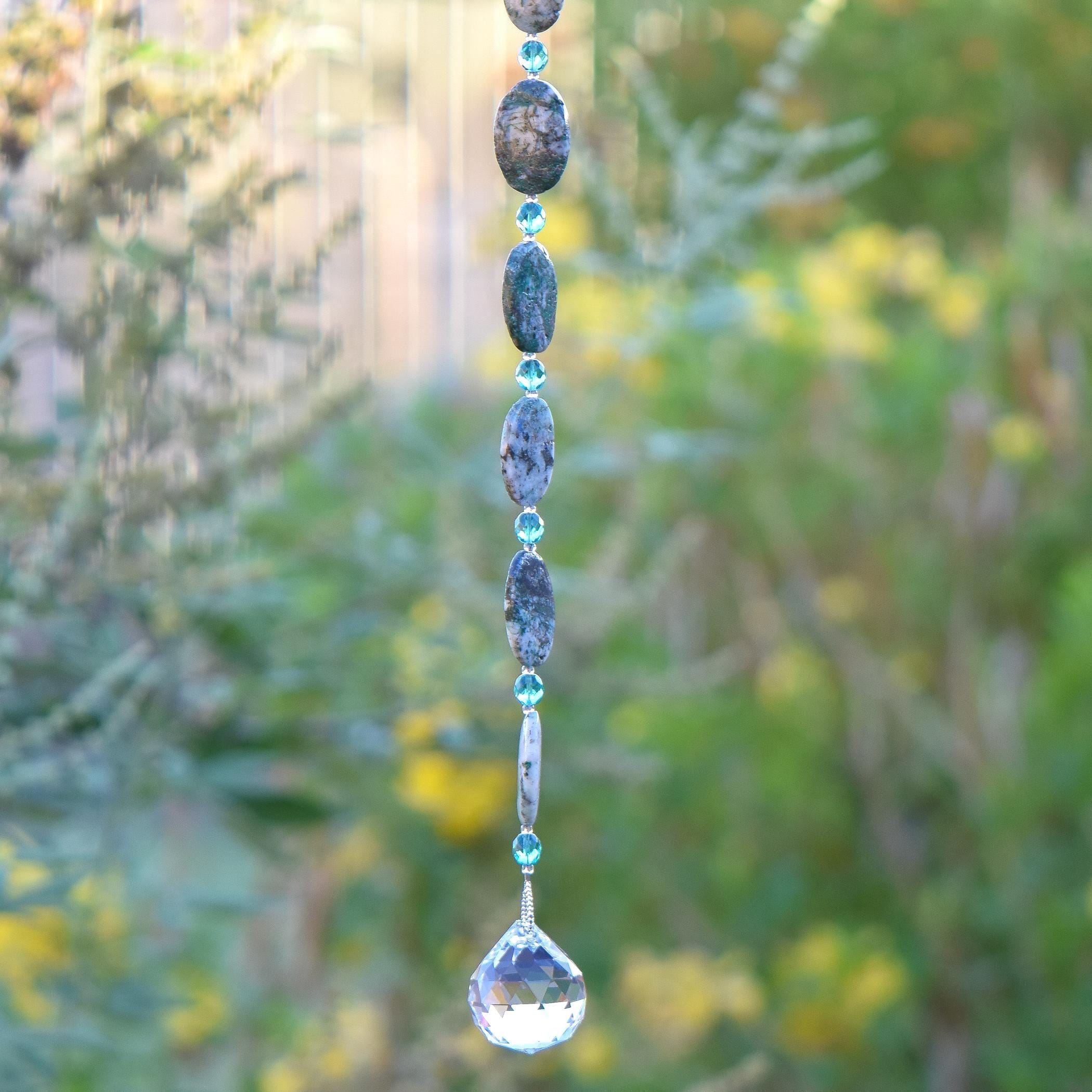 Long hanging sun catcher made of large oval azurite malachite stone beads paired with small turquoise glass beads and a large crystal prism. This photo shows the bottom 5 stone beads, 6 glass beads and the prism, against a blurred foliage background.