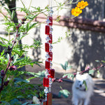 Two ends of a long stainless steel wire, hanging vertically with handmade fused glass rectangular beads in red, paired with clear glass beads. Each end of the wire is anchored by a long rectangular piece of red fused glass. The chime is hanging in a garden with yellow cosmos flowers and a small white dog in the background.