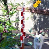 Two ends of a long stainless steel wire, hanging vertically with handmade fused glass rectangular beads in red, paired with clear glass beads. Each end of the wire is anchored by a long rectangular piece of red fused glass. The chime is hanging in a garden with yellow cosmos flowers and a small white dog in the background.