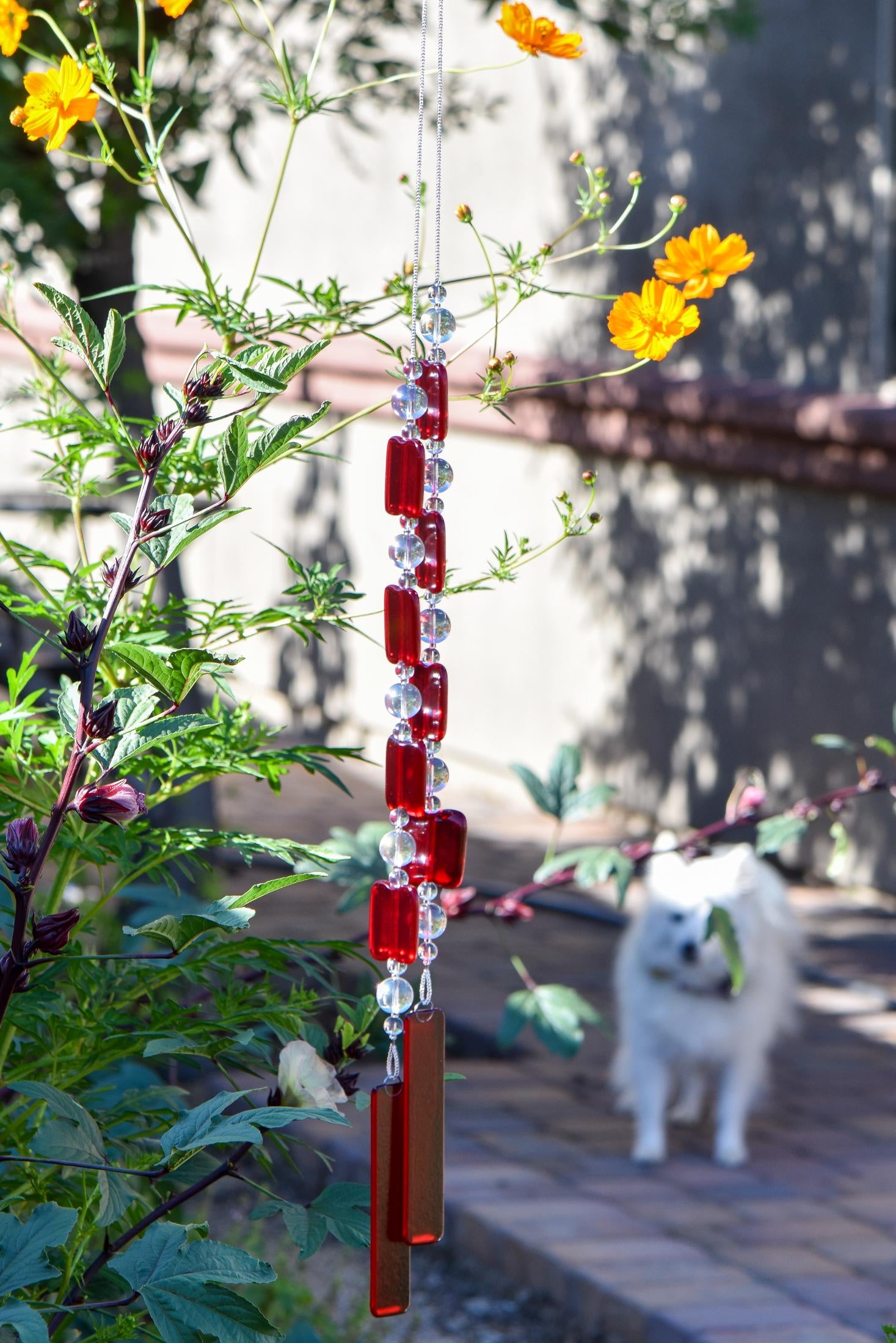 Two ends of a long stainless steel wire, hanging vertically with handmade fused glass rectangular beads in red, paired with clear glass beads. Each end of the wire is anchored by a long rectangular piece of red fused glass. The chime is hanging in a garden with yellow cosmos flowers and a small white dog in the background.