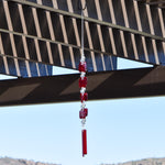 Two ends of a long stainless steel wire, hanging vertically with handmade fused glass rectangular beads in red, paired with clear glass beads. Each end of the wire is anchored by a long rectangular piece of red fused glass. Chime is hanging from a hook that is hanging from a pergola. View is looking up with sky and a mountain in the background.