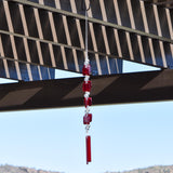 Two ends of a long stainless steel wire, hanging vertically with handmade fused glass rectangular beads in red, paired with clear glass beads. Each end of the wire is anchored by a long rectangular piece of red fused glass. Chime is hanging from a hook that is hanging from a pergola. View is looking up with sky and a mountain in the background.