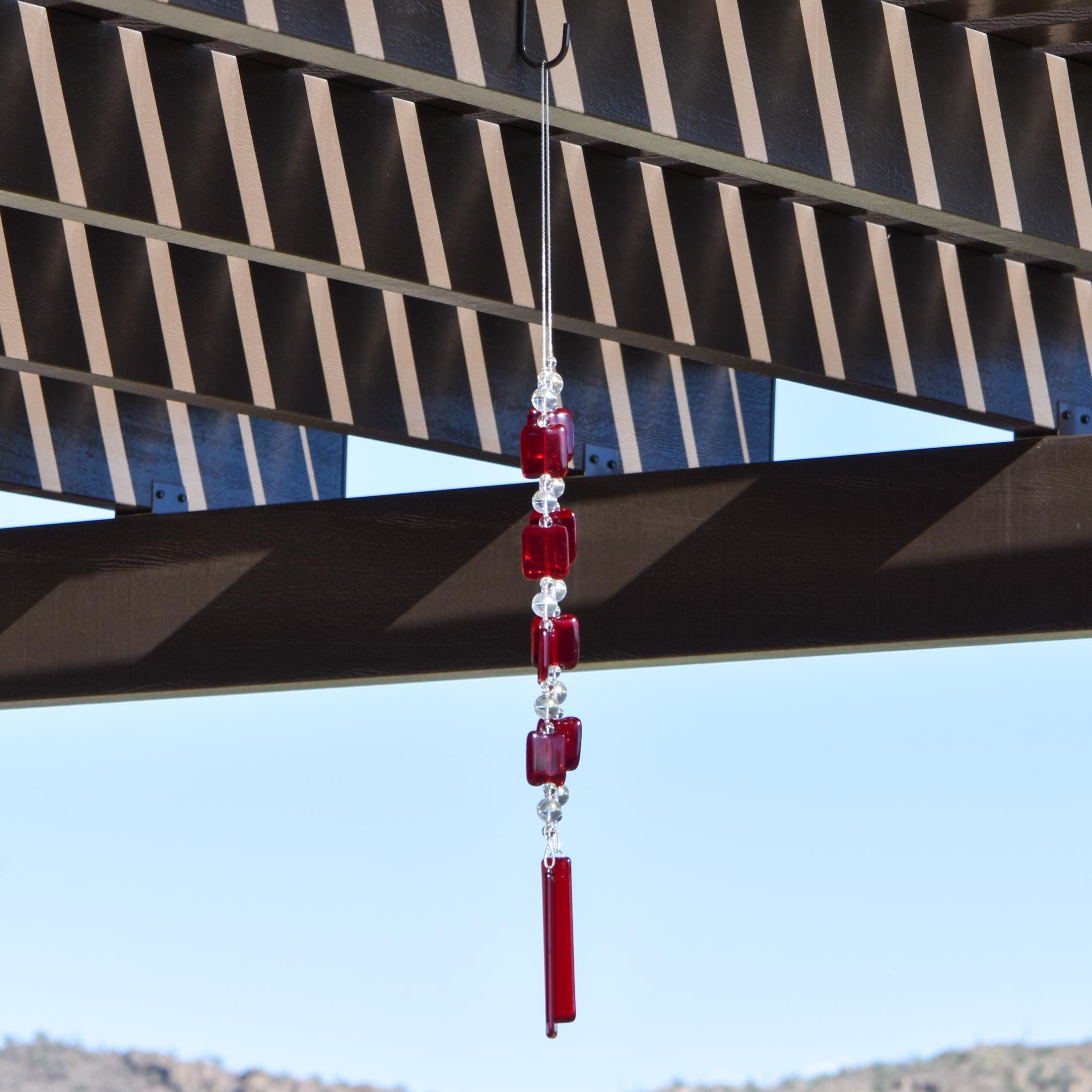 Two ends of a long stainless steel wire, hanging vertically with handmade fused glass rectangular beads in red, paired with clear glass beads. Each end of the wire is anchored by a long rectangular piece of red fused glass. Chime is hanging from a hook that is hanging from a pergola. View is looking up with sky and a mountain in the background.