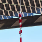 Two ends of a long stainless steel wire, hanging vertically with handmade fused glass rectangular beads in red, paired with clear glass beads. Each end of the wire is anchored by a long rectangular piece of red fused glass. Chime is hanging from a hook that is hanging from a pergola. View is looking up with sky and a mountain in the background.