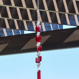 Two ends of a long stainless steel wire, hanging vertically with handmade fused glass rectangular beads in red, paired with clear glass beads. Each end of the wire is anchored by a long rectangular piece of red fused glass. Chime is hanging from a hook that is hanging from a pergola. View is looking up with sky and a mountain in the background.