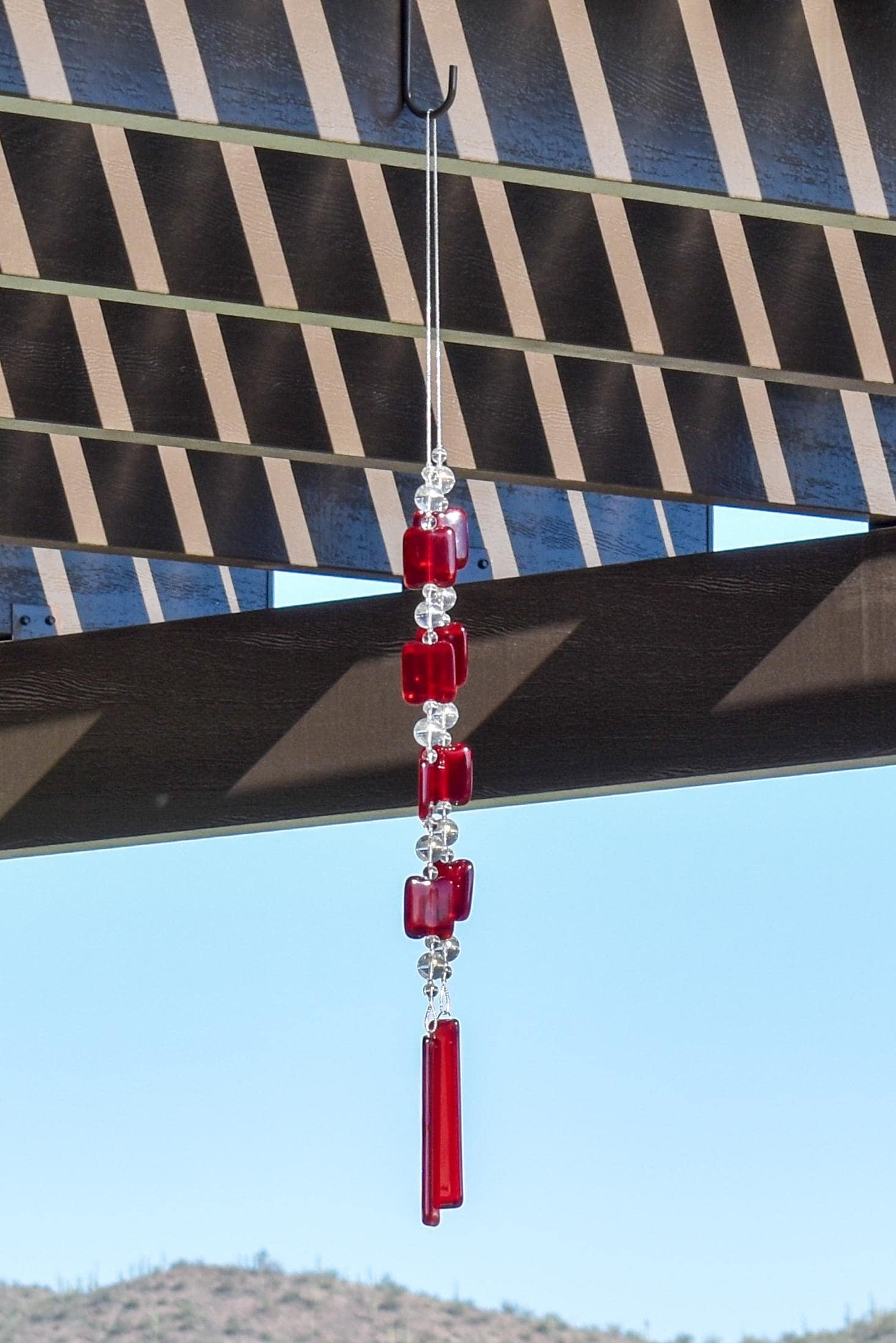 Two ends of a long stainless steel wire, hanging vertically with handmade fused glass rectangular beads in red, paired with clear glass beads. Each end of the wire is anchored by a long rectangular piece of red fused glass. Chime is hanging from a hook that is hanging from a pergola. View is looking up with sky and a mountain in the background.