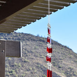 Two ends of a long stainless steel wire, hanging vertically with handmade fused glass rectangular beads in red, paired with clear glass beads. Each end of the wire is anchored by a long rectangular piece of red fused glass. Chime is hanging from a hook that is hanging from a pergola. View is looking up with sky and a mountain in the background.