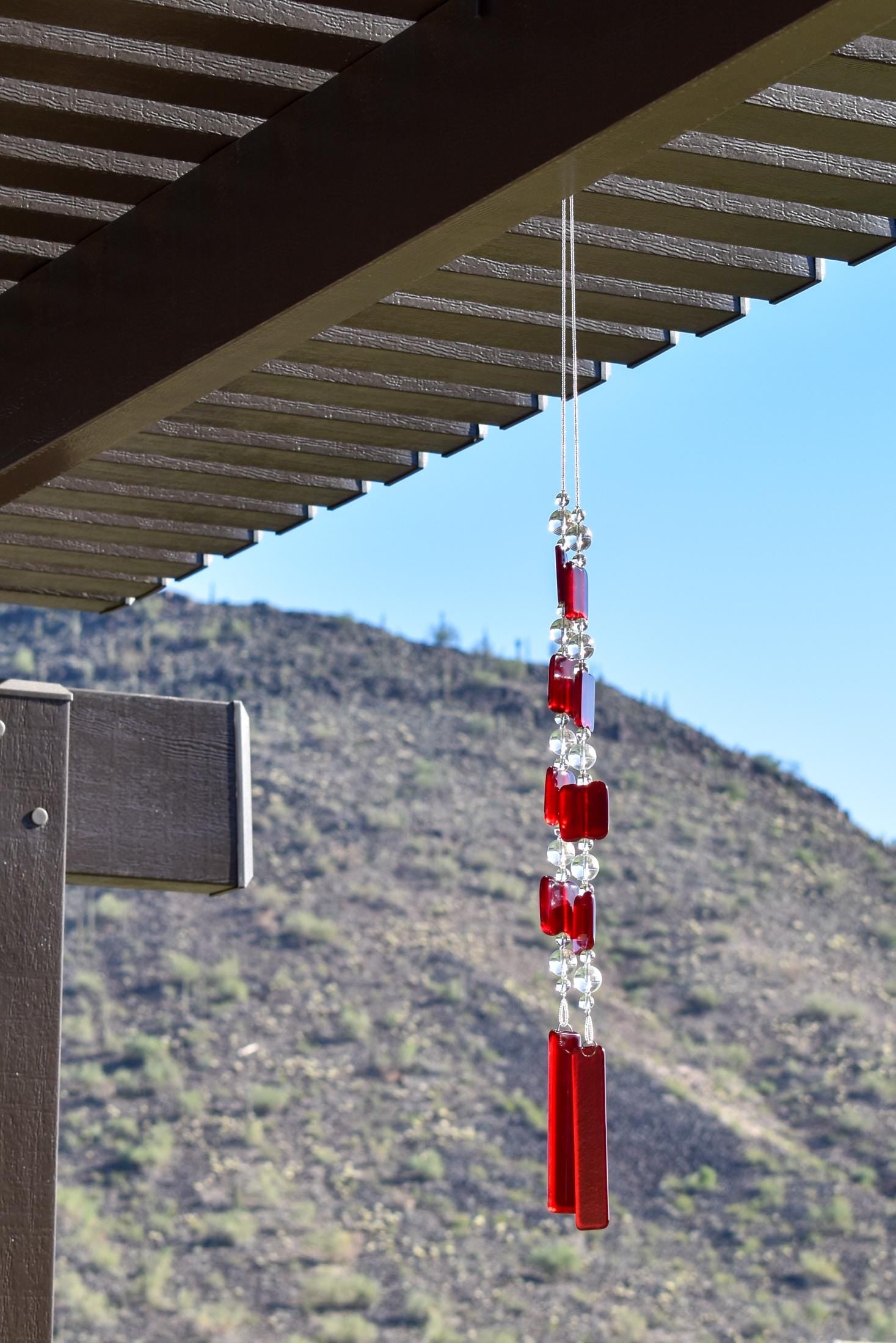Two ends of a long stainless steel wire, hanging vertically with handmade fused glass rectangular beads in red, paired with clear glass beads. Each end of the wire is anchored by a long rectangular piece of red fused glass. Chime is hanging from a hook that is hanging from a pergola. View is looking up with sky and a mountain in the background.