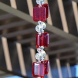 Close up view of handmade fused glass beads, rectangularly shaped and flat. Strung with clear glass beads with pergola in the background.