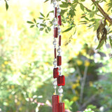Two ends of a long stainless steel wire, hanging vertically from a tree with handmade fused glass rectangular beads in red, paired with clear glass beads. Each end of the wire is anchored by a long rectangular piece of red fused glass. Blurred foliage in background.