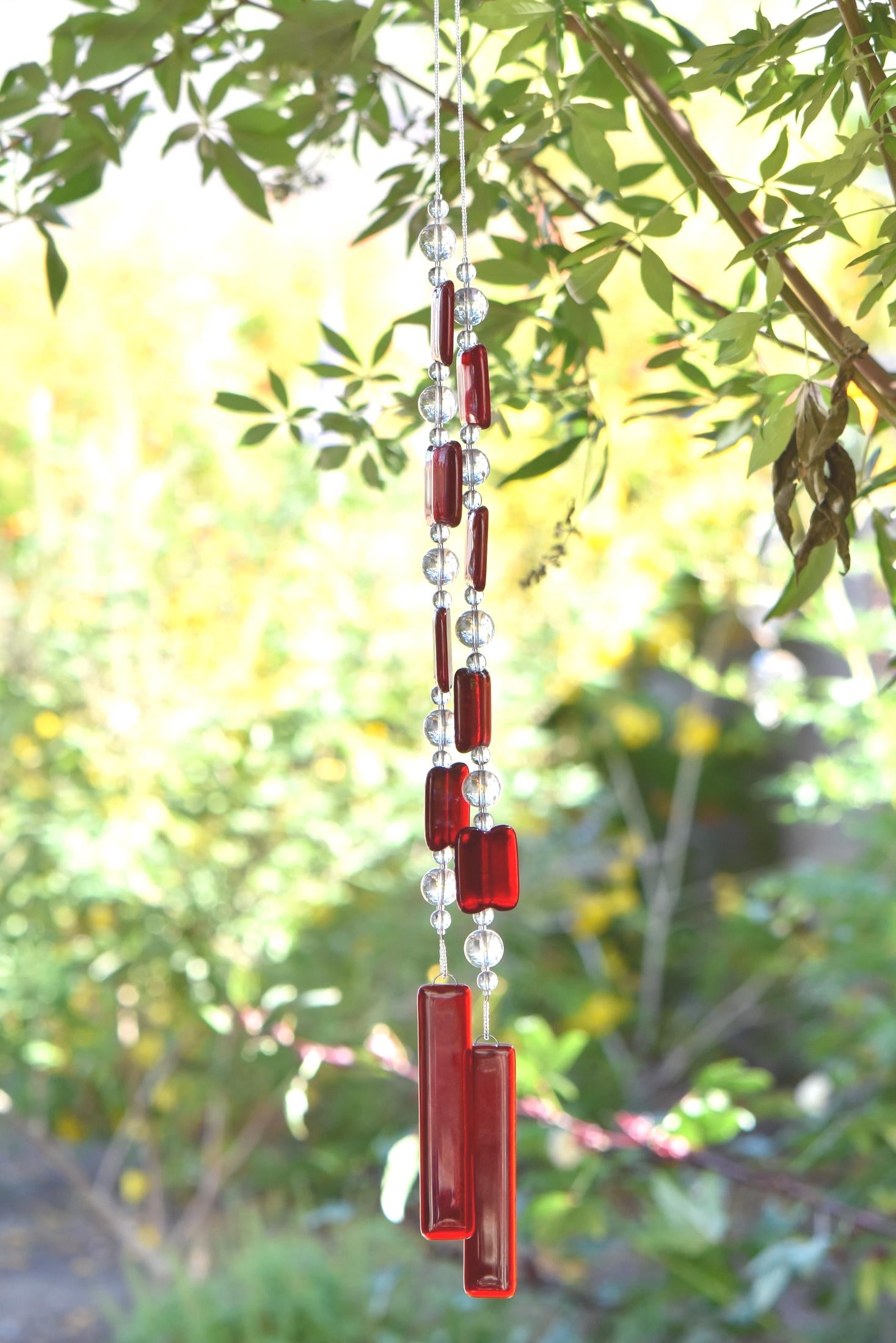 Two ends of a long stainless steel wire, hanging vertically from a tree with handmade fused glass rectangular beads in red, paired with clear glass beads. Each end of the wire is anchored by a long rectangular piece of red fused glass. Blurred foliage in background.