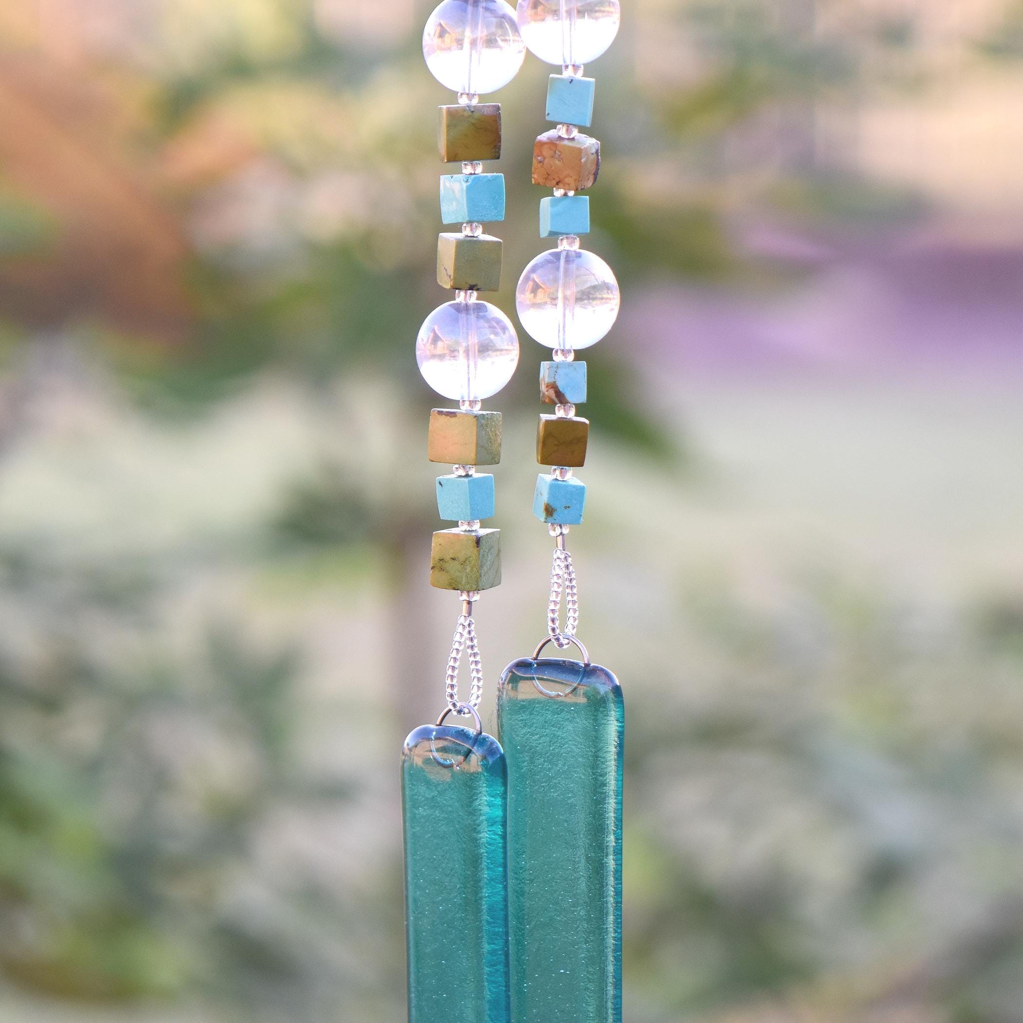 Two ends of a long wire hanging down covered in beads with glass anchors. The beads include cubic stones of howlite or magnesite, large clear glass beads with a reflective finish and glass silver-lined seed beads. The anchor glass is kiln formed, caribbean blue transparent. Background is blurred foliage.