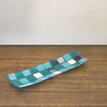 A rectangular glass tray with a mosaic tile design. The tray features a pattern of square tiles in shades of teal and white. The tiles are arranged in a grid-like pattern, creating a visually appealing surface. The tray is a wood surface with white in the background.