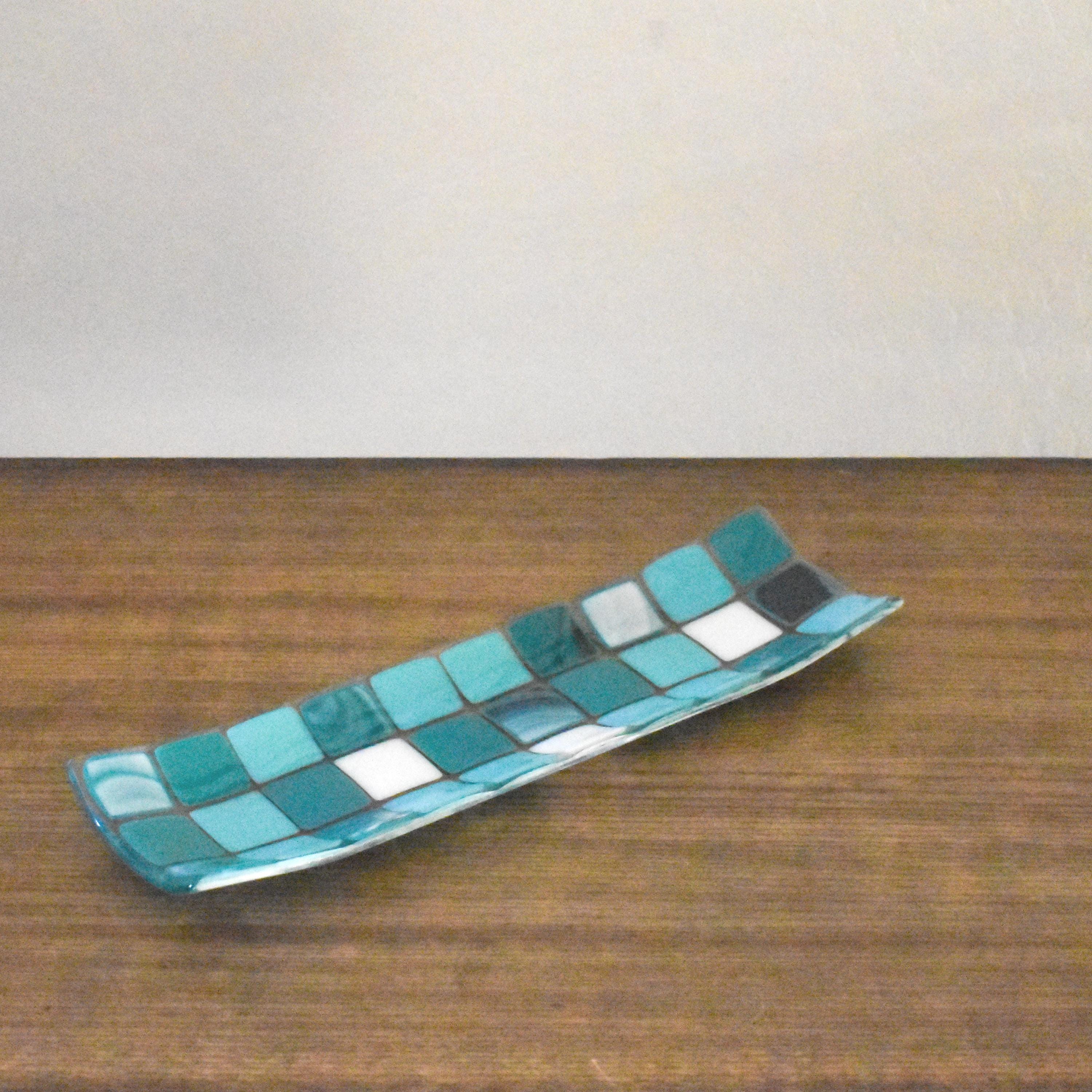 A rectangular glass tray with a mosaic tile design. The tray features a pattern of square tiles in shades of teal and white. The tiles are arranged in a grid-like pattern, creating a visually appealing surface. The tray is a wood surface with white in the background.