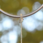 Close-up view of a silver metal wire covered with silver-lined seed beads and attached to a circular metal rod. The background is blurred, suggesting an outdoor setting with bokeh effects of green and white.
