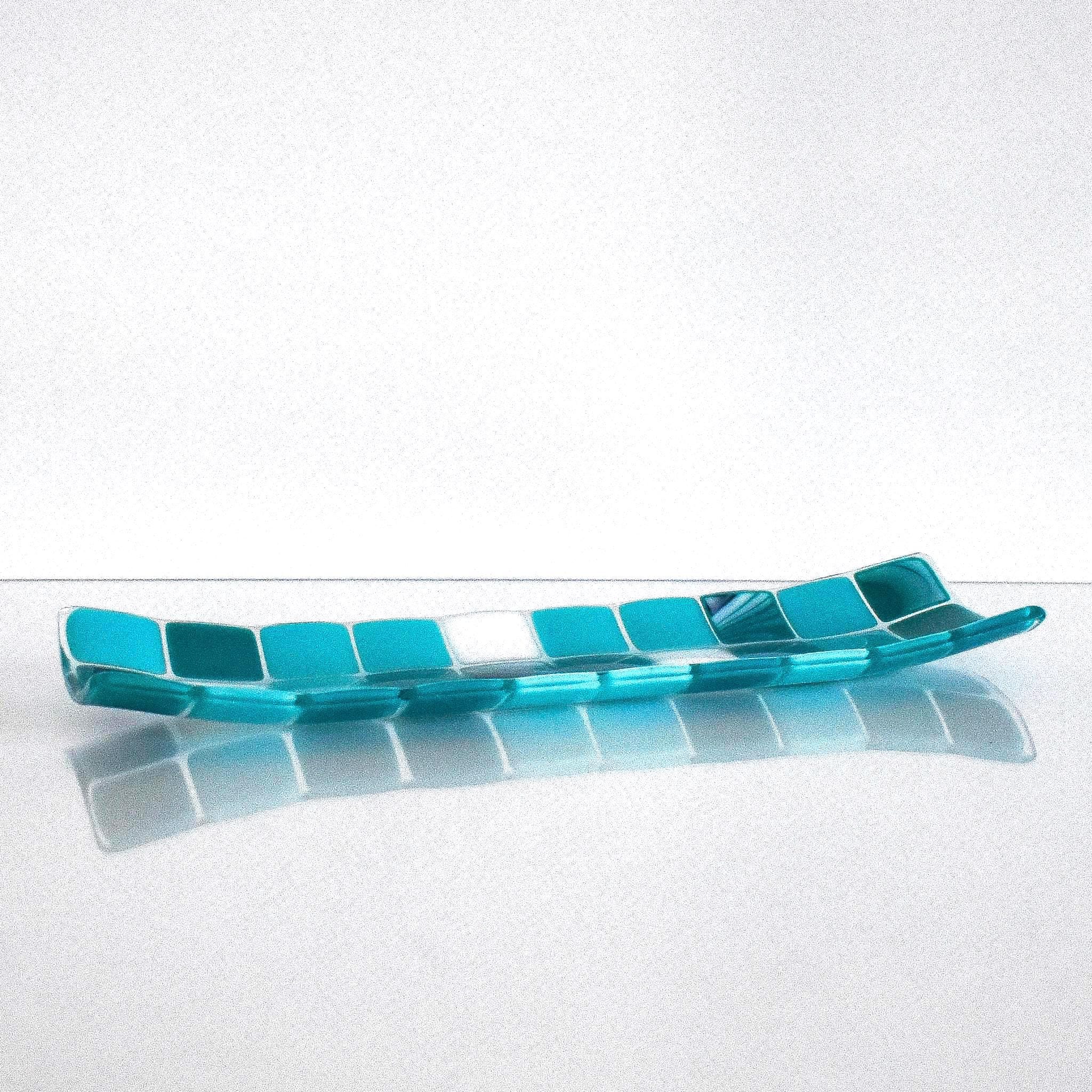 A rectangular glass tray with a mosaic tile design. The tray features a pattern of square tiles in shades of teal and white. The tiles are arranged in a grid-like pattern, creating a visually appealing surface. The tray is placed on a white surface.