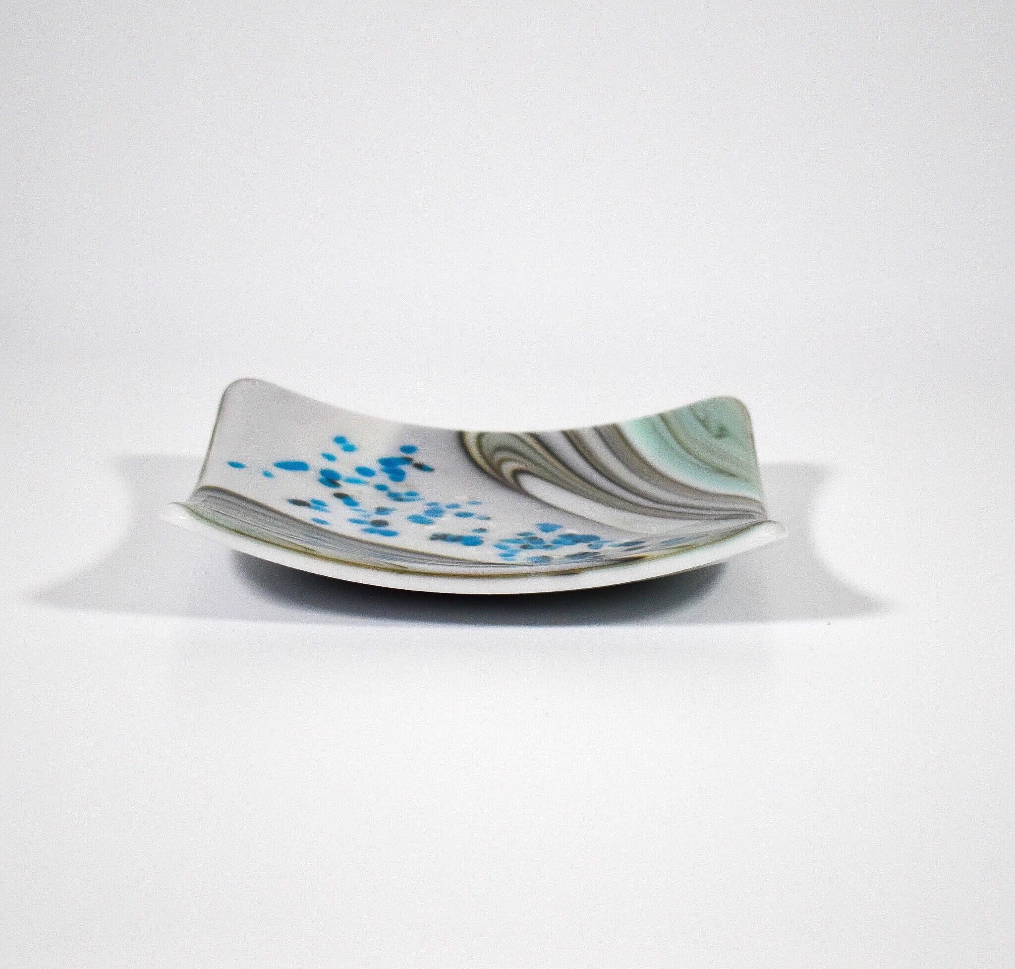 Six inch square glass tray with modern design on wood table.Tray is white with swirls and flakes of aqua, cream, tan and gray. On a white surface. Viewed from the side.