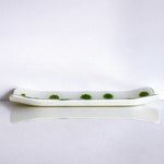 A long, rectangular dish with a curved shape. The dish is white with a series of green, circular designs evenly spaced along the top surface. The dish is resting on a white reflective surface, creating a mirrored effect.