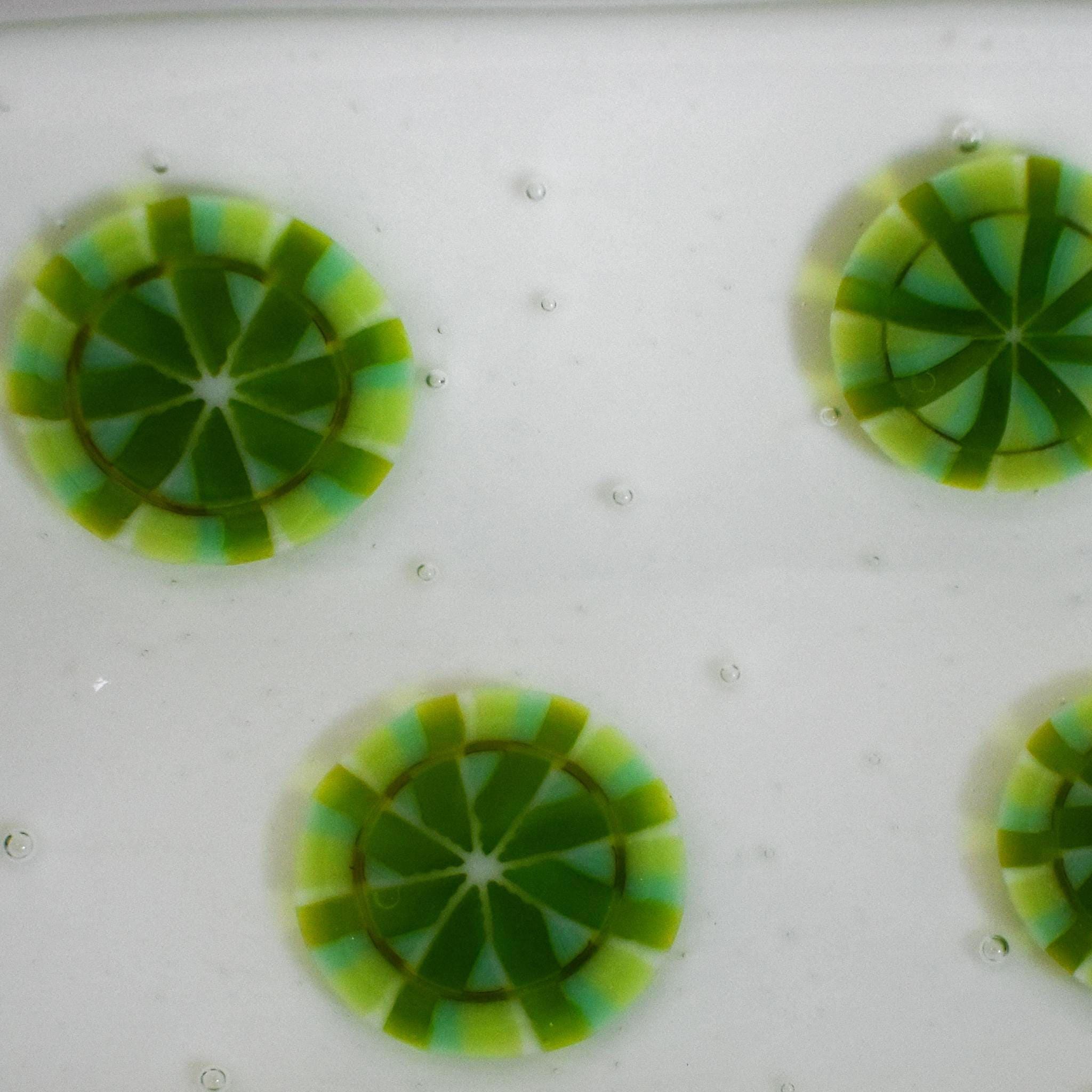 Close-up of a white surface with four circular, decorative glass pieces. Each piece features a lime green and light green segmented design, resembling a citrus fruit slice. Small, clear bubbles are scattered across the white background, adding texture.