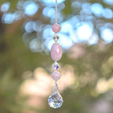 Large oval rose quartz bead flanked by Austrian crystal and two smaller round rose quartz beads, hanging vertically from a tree, anchored by round crystal prism.