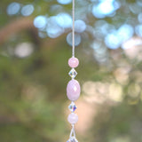 Large oval rose quartz bead flanked by Austrian crystal and two smaller round rose quartz beads, hanging vertically from a tree, anchored by round crystal prism.