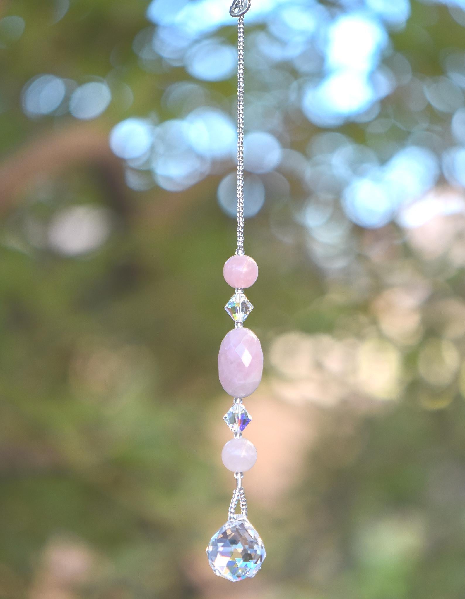 Large oval rose quartz bead flanked by Austrian crystal and two smaller round rose quartz beads, hanging vertically from a tree, anchored by round crystal prism.