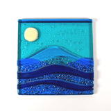 Six inch square glass panel on a clear acrylic flat on a white surface. The panel depicts waves of blue with texture and flow underneath a full moon in an evening sky.