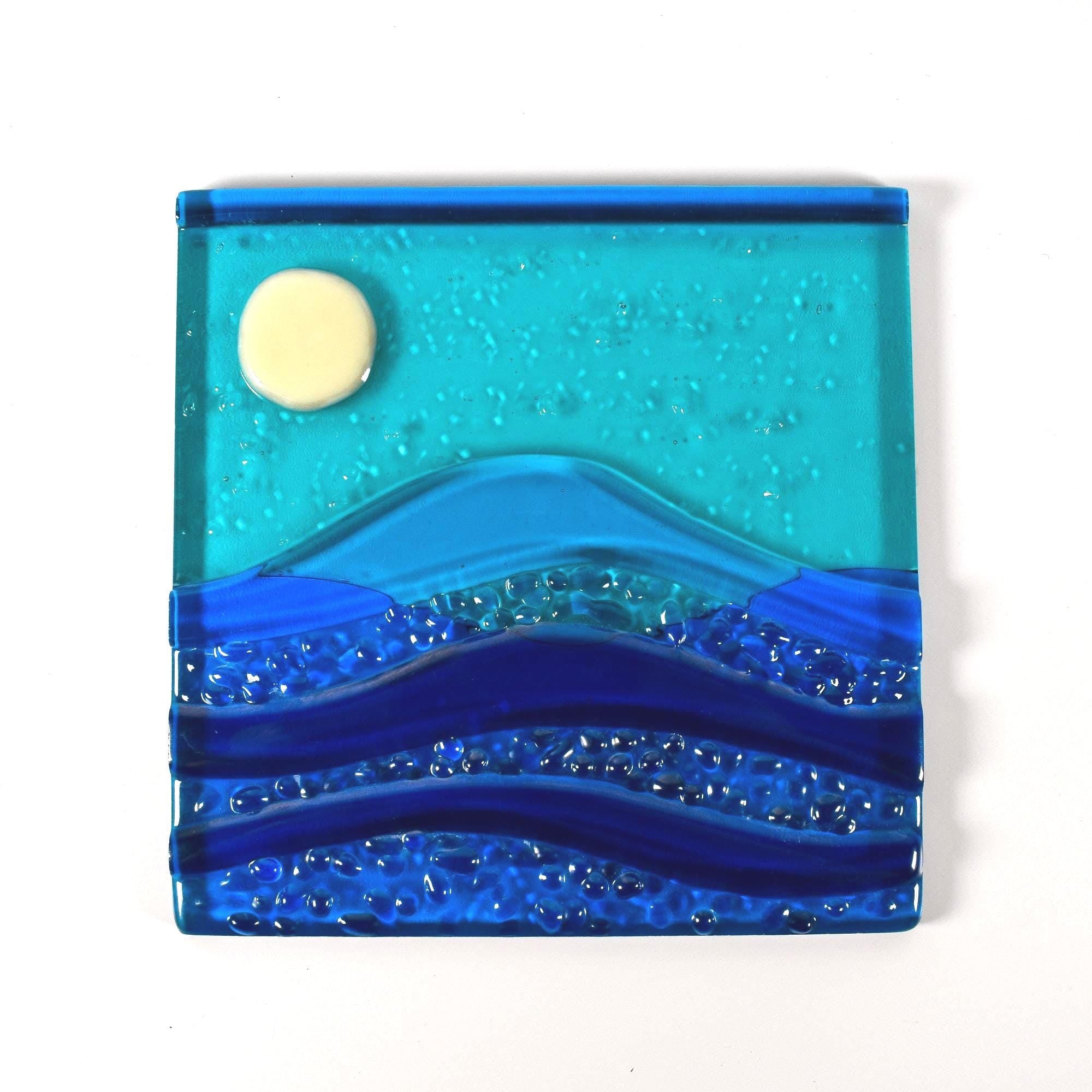 Six inch square glass panel on a clear acrylic flat on a white surface. The panel depicts waves of blue with texture and flow underneath a full moon in an evening sky.