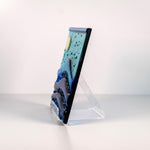 Six inch square glass panel on a clear acrylic stand, viewed from the side showing the edge of the piece and the back of the acrylic easel stand. The surface and background are white. The panel depicts waves of blue with texture and flow underneath a full moon in an evening sky.