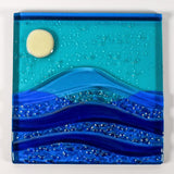 Six inch square glass panel flat on a white surface. The panel depicts waves of blue with texture and flow underneath a full moon in an evening sky.
