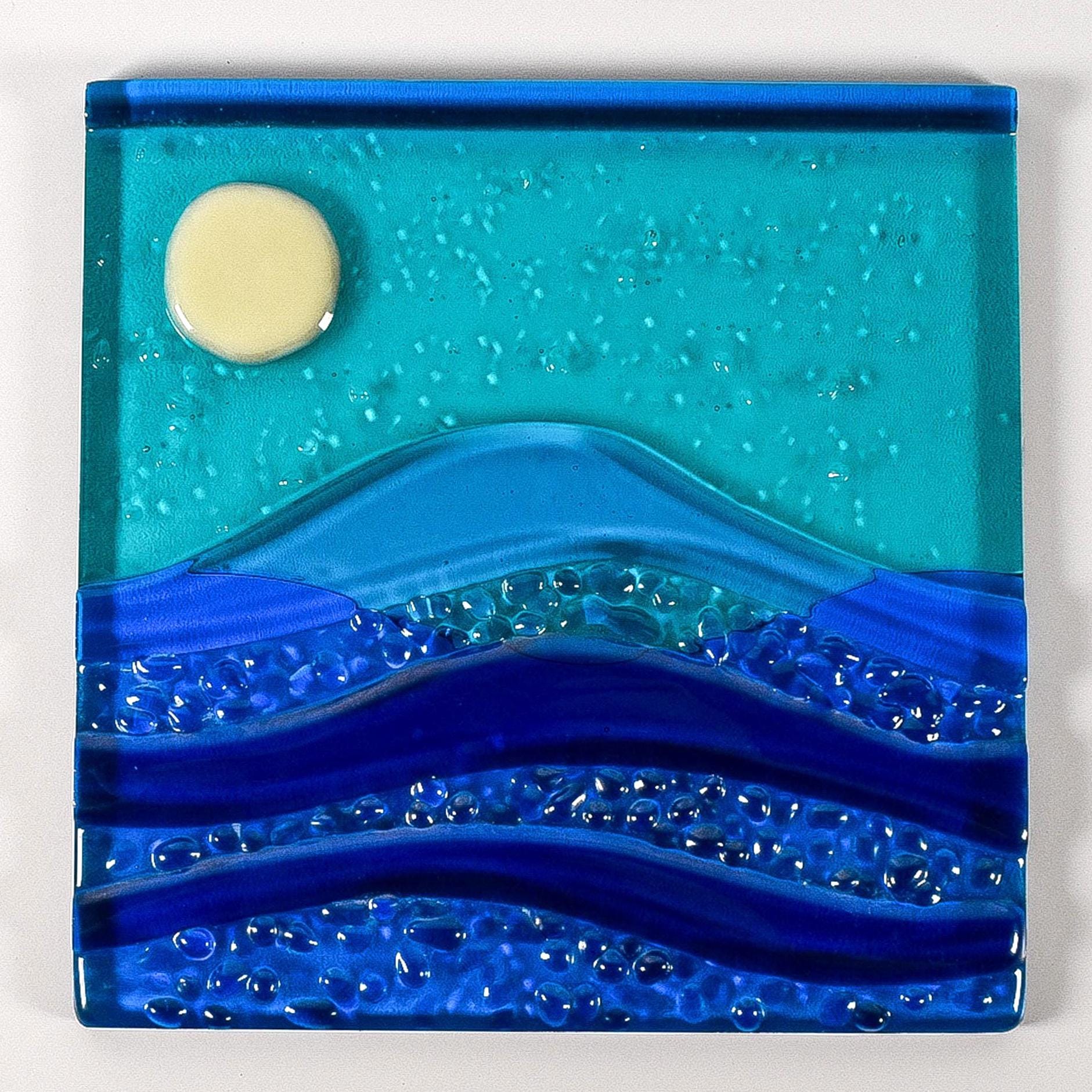 Six inch square glass panel flat on a white surface. The panel depicts waves of blue with texture and flow underneath a full moon in an evening sky.