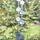 Bottom of hanging wind chime from Cinjo Handmade. Six large sea glass beads are pictured, strung with smaller clear glass beads and anchored by two pieces of kiln formed glass. The chime is hanging in front of blurred foliage.
