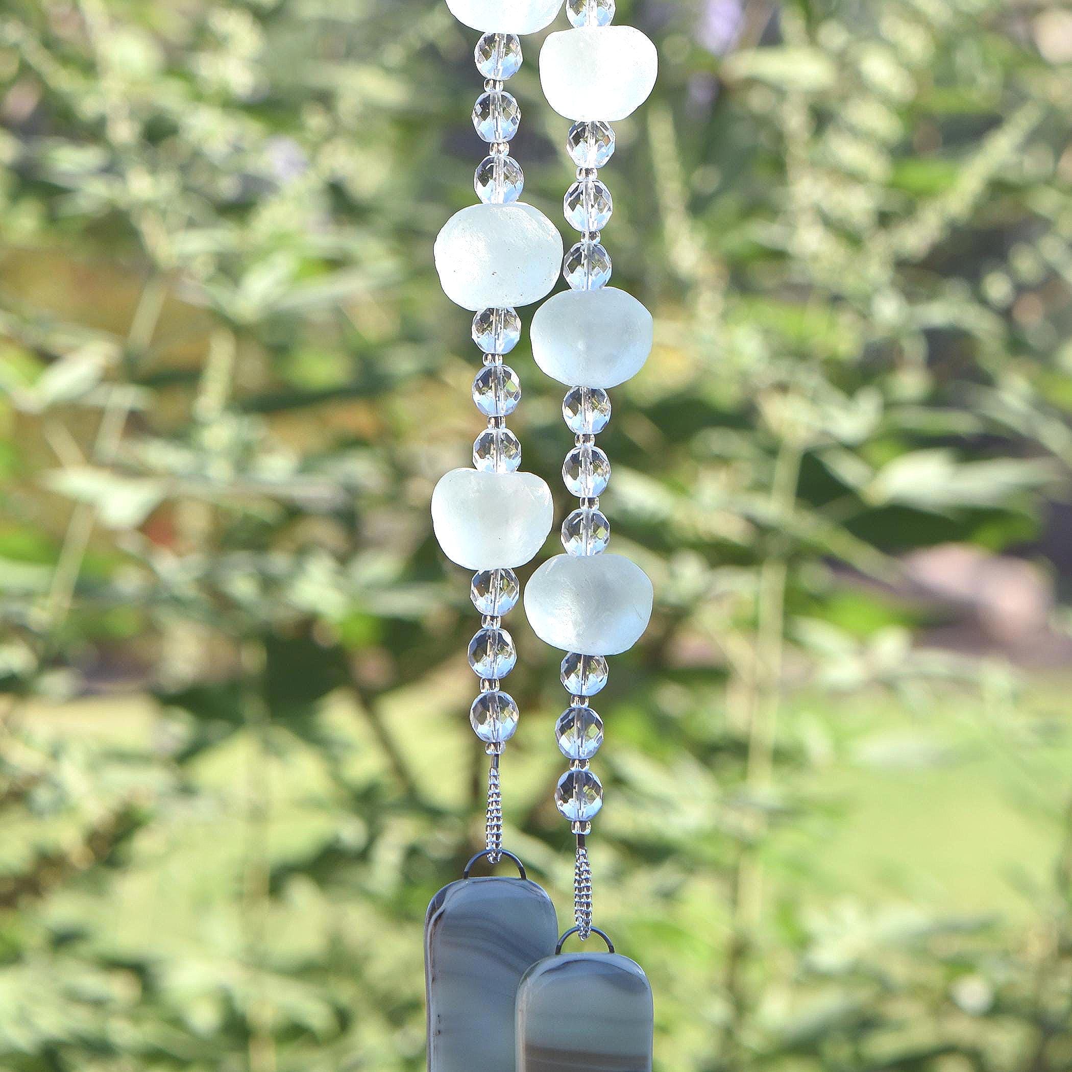 Bottom of hanging wind chime from Cinjo Handmade. Six large sea glass beads are pictured, strung with smaller clear glass beads and anchored by two pieces of kiln formed glass. The chime is hanging in front of blurred foliage.