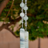 Bottom of hanging wind chime from Cinjo Handmade. Four large sea glass beads are pictured, strung with smaller clear glass beads and anchored by two pieces of kiln formed glass. The chime is hanging in front of blurred foliage.