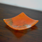 Square fused glass tray made of clear glass with streaks of bright orange, viewed from an agle as it is sitting on a dark brown wood table.