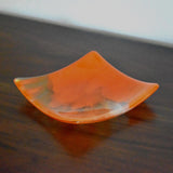 Square fused glass tray made of clear glass with streaks of bright orange, viewed from an agle as it is sitting on a dark brown wood table.