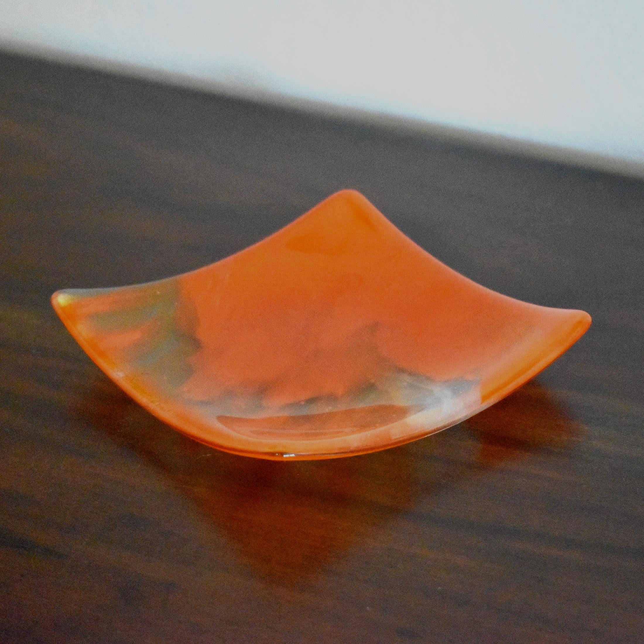 Square fused glass tray made of clear glass with streaks of bright orange, viewed from an agle as it is sitting on a dark brown wood table.