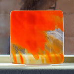 Fused glass tray made of clear glass with streaks of bright orange. Sitting on stand on window sill with tree trunk behind.