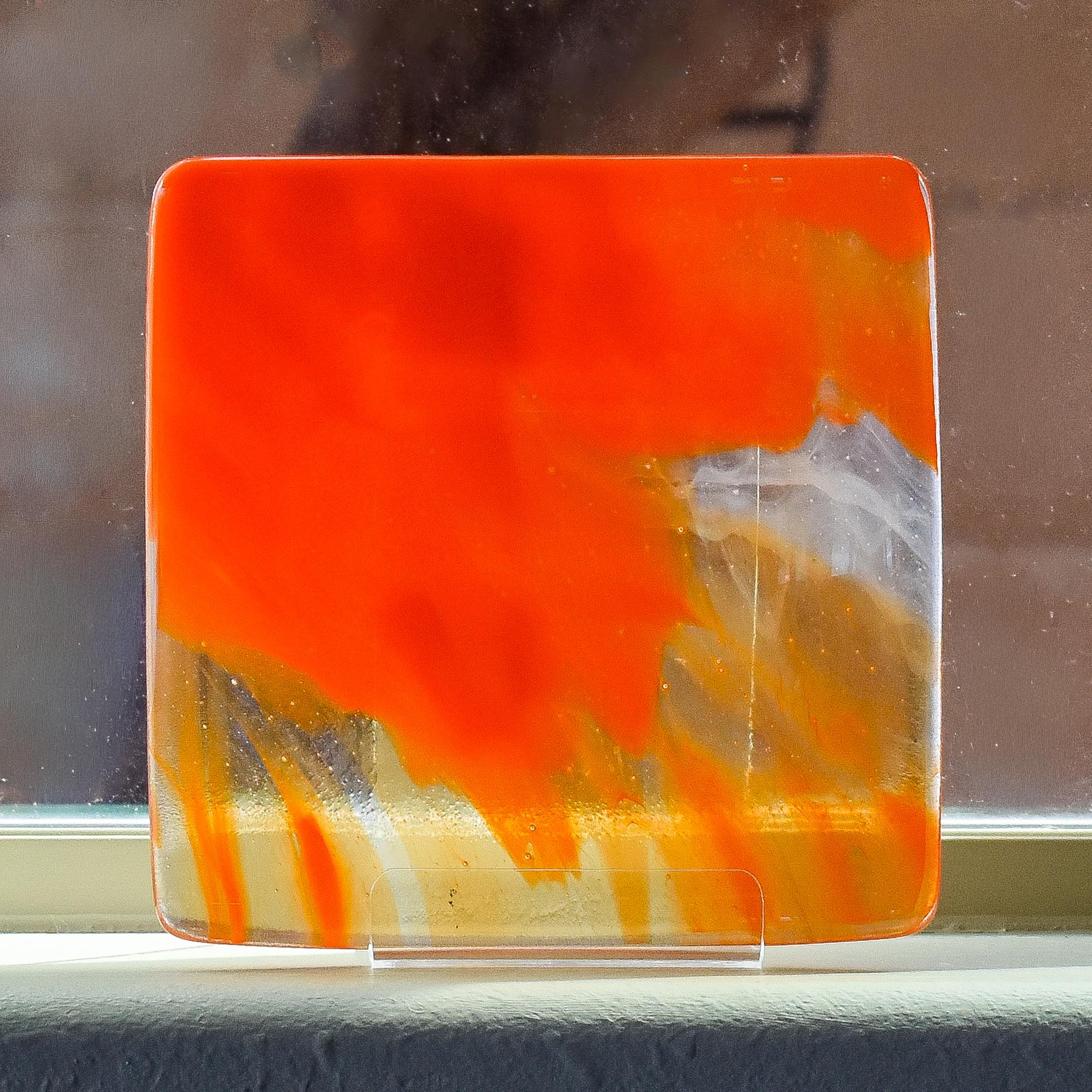 Fused glass tray made of clear glass with streaks of bright orange. Sitting on stand on window sill with tree trunk behind.