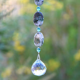 The bottom of a long hanging sun catcher made of large oval azurite malachite stone beads paired with small turquoise glass beads and a large crystal prism. This photo shows the bottom 3 stone beads, 3 glass beads and the prism, against a blurred foliage background.