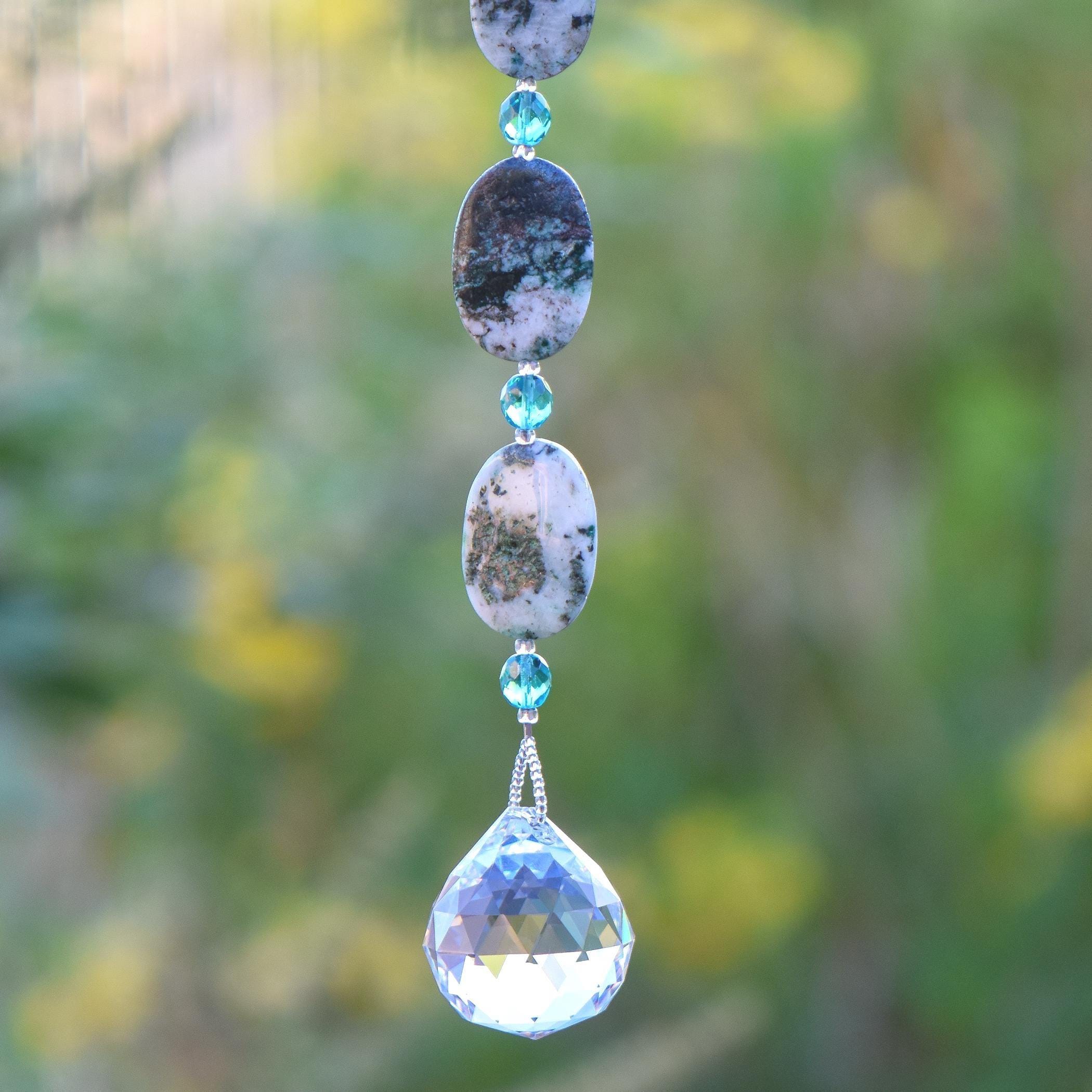 The bottom of a long hanging sun catcher made of large oval azurite malachite stone beads paired with small turquoise glass beads and a large crystal prism. This photo shows the bottom 3 stone beads, 3 glass beads and the prism, against a blurred foliage background.